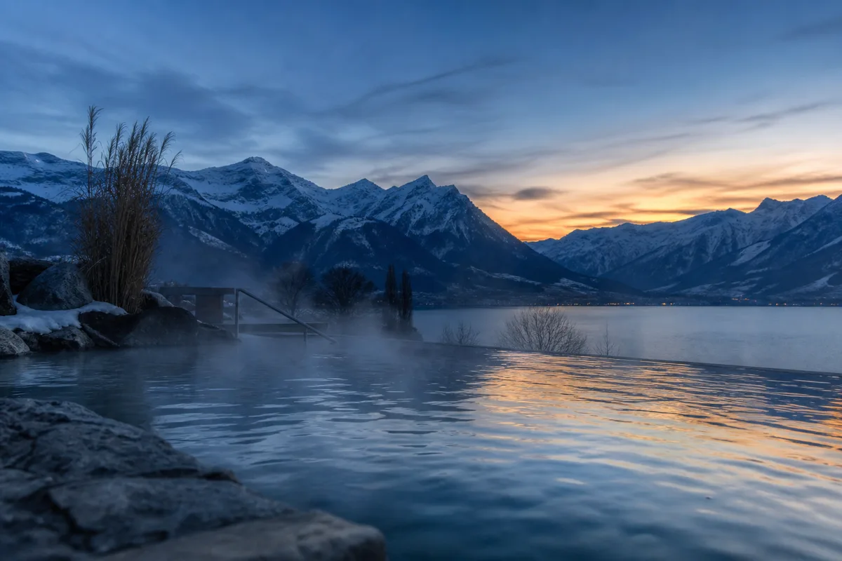 Bassin thermal extérieur fumant devant un massif alpin enneigé au crépuscule